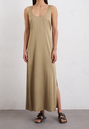 Vrouw draagt een beige mouwloze maxi-jurk met zijsplit, gecombineerd met zwarte sandalen met gesp, staande tegen een lichte gestructureerde muur.