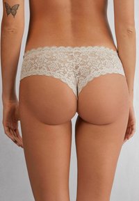 Lace bikini ondergoed in lichtbeige met een gegolfde rand. Bevat bloem- en geometrische patronen. Zachte textuur met minimale bedekking.
