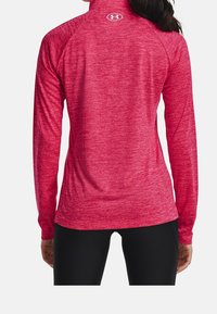 Kvinna iklädd en åtsittande röd långärmad träningströja med en vit Under Armour-logotyp på den övre ryggen, matchad med svarta leggings.