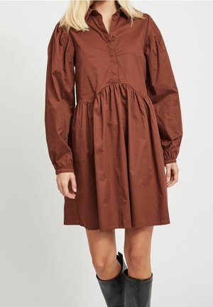 Femme portant une robe marron à manches longues et boutonnée, avec une taille froncée, et des bottes noires montant jusqu'aux genoux, debout devant un fond uni.