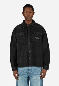 Czarna kurtka kordowa z dwoma kieszeniami na piersi, zapięciem na guziki i haftowanym logo z przodu. Luźny krój z lekko teksturowanym materiałem.