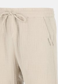 Pantalons beiges texturés avec une taille élastique, une fermeture à cordon de serrage et des poches latérales. Présente une marque subtile sur la poche.
