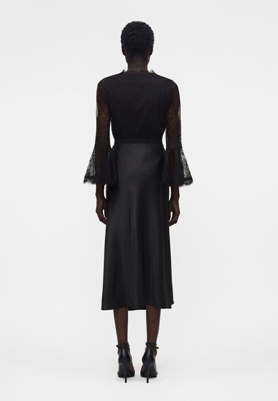 Γυναίκα φορώντας μαύρο μίντι φόρεμα με διαφανή δαντελένια μακριά μανίκια με καμπάνα στα μανσέτα, συνδυασμένο με μαύρα ψηλοτάκουνα παπούτσια με λουράκι στον αστράγαλο.