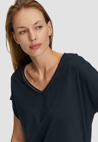 Vrouw met lichtbruin haar die een marineblauwe V-hals shirt met gedetailleerde stiksels draagt, kijkt naar links tegen een effen lichte achtergrond.