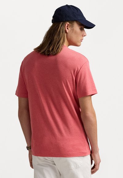 Tricou roșu cu mâneci scurte, dintr-un material moale și texturat, cu decolteu rotund și o croială lejeră, parțial ascuns de corpul purtătorului.