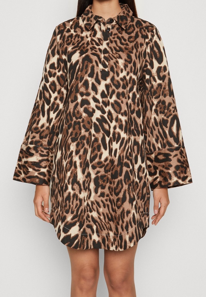 Femme portant une robe mi-longue à manches longues avec un imprimé léopard abstrait marron, beige et noir et un petit col avec fermeture par bouton.