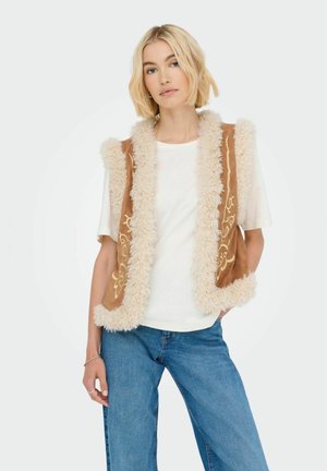 Braune Kunstsuede-Weste mit beigem, flauschigem Besatz und floralem Stickerei auf der Vorderseite, getragen über einem weißen Hemd und kombiniert mit blauen Jeans.