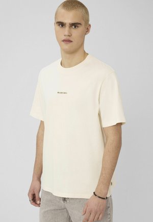 Jeune homme aux cheveux courts décolorés portant un T-shirt crème à manches courtes avec le texte "BAROUDEUR" et un jean gris clair, debout devant un fond uni.