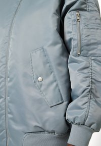 Giacca bomber blu chiaro con una finitura liscia e lucida, polsini a coste, una tasca laterale con zip e un pulsante a scatto sulla tasca frontale.