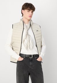 Světlý béžový prošívaný vest s vysokým límcem, černým lemováním, nošený přes bílou košili s dlouhým rukávem, v kombinaci s tmavými džíny a vzorovaným šátkem.