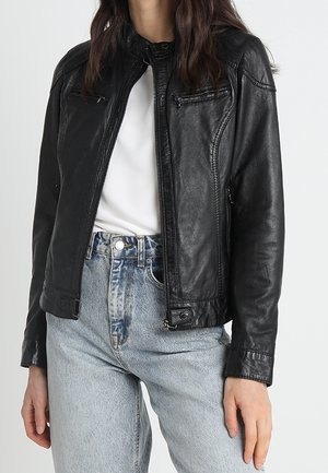 Femme portant une veste en cuir noire zippée sur une chemise blanche et un jean taille haute bleu clair, debout avec une main détendue sur le côté.