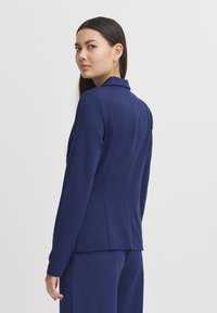 Chaqueta ajustada de color azul marino en una tela suave, con un cuello estructurado, forma entallada y mangas largas, que presenta costuras limpias y un minimalismo en los detalles.