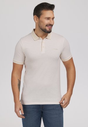 Mężczyzna z brodą ubrany w jasnobeżową koszulkę polo z krótkim rękawem i niebieskie dżinsy, uśmiechający się i patrzący w prawo na jednolitym tle.