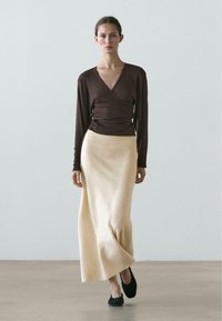 Brun langærmet omslagstop med dyb V-hals, kombineret med en cremefarvet, ankel-lang nederdel. Sorte flade sko fuldender outfittet.