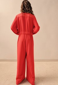 Combinaison rouge à manches longues, taille élastique et jambes larges. Tissu lisse avec des coutures minimales, vue de dos sur un fond neutre.