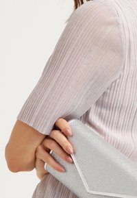 Pochette à paillettes argentées avec un rabat triangulaire épuré, tenue contre une manche côtelée mauve clair, mettant en valeur un éclat subtil et une texture lisse.