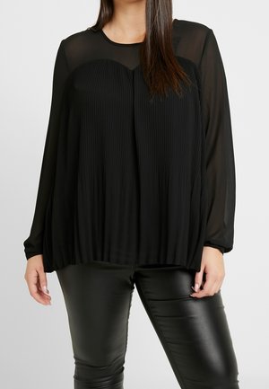 Kvinde iført en sort, gennemsigtig bluse med lange ærmer og plisserede detaljer foran kombineret med sorte bukser med læderlook.