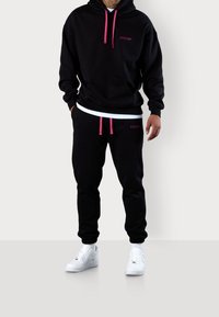 Schwarzes Hoodie- und Jogginganzug-Set mit pinken Kordeln und Akzenten, ausgestattet mit einer Kängurutasche und gerippten Bündchen, kombiniert mit weißen Turnschuhen.