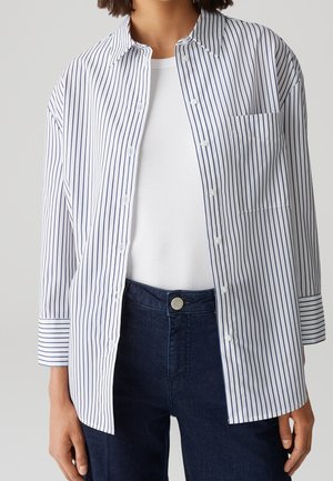 Osoba ubrana w białą koszulę zapinaną na guziki z cienkimi pionowymi granatowymi paskami, pod którą ma białą prążkowaną bluzkę oraz ciemnoniebieskie dżinsy.