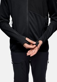 Giacca nera con zip e pannello testurizzato, con aperture per il pollice, indossata su pantaloni scuri con tasche con zip, che mostrano caratteristiche di design regolabili.
