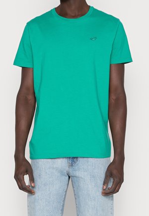 Grünes Baumwoll-T-Shirt mit Rundhalsausschnitt, kurzen Ärmeln und einem kleinen Logo-Akzent auf der linken Brust. Getragen mit hellblauen Jeans.