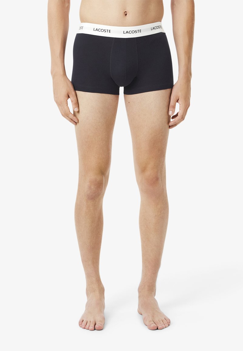 Schwarze Boxershorts aus elastischem Stoff, mit einem weißen Lacoste-Logo-Bund, eng anliegender Passform und Oberschenkelabdeckung. Glatte Textur.