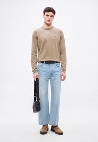 Beżowa koszula z długim rękawem z logo, jasnoniebieskie szerokie spodnie dżinsowe, czarny pasek, brązowe zamszowe buty i czarna torebka na ramię. Jednolite tło.