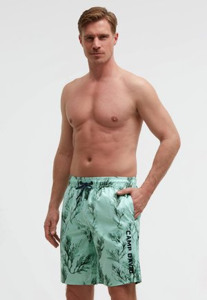 Oben ohne stehender Mann, der hellgrüne Badeshorts mit schwarzem Seetangmuster und dem Schriftzug "CAMP DAVID" trägt, vor einem schlichten Hintergrund.
