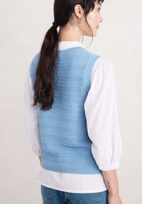 Gilet tricoté bleu clair avec des motifs texturés verticaux, porté sur une blouse blanche à manches bouffantes. Complété par un jean en denim.