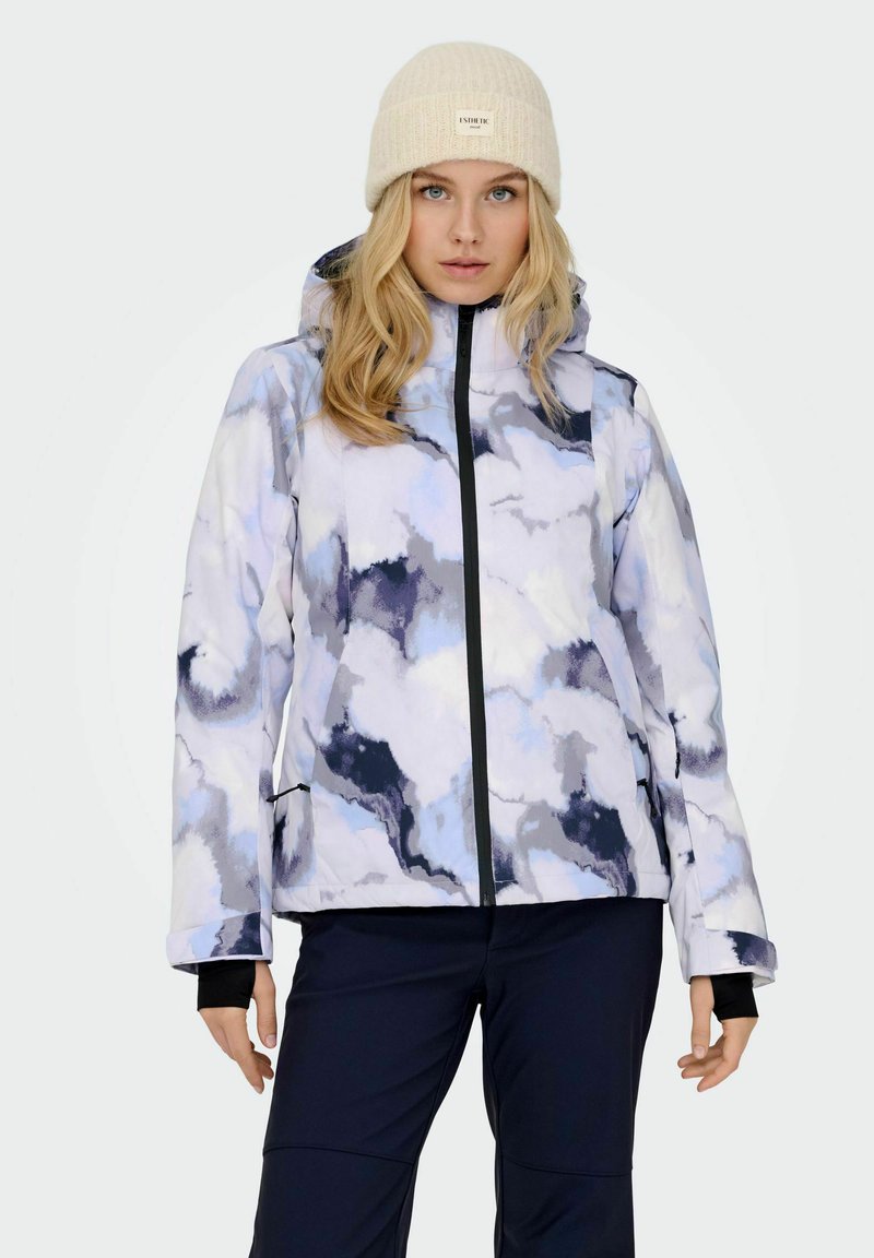 Kvinde iført en lys tie-dye vinterjakke med hætte, sorte bukser og en beige strikket beanie, stående foran en ensfarvet baggrund.