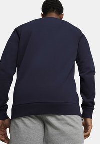 Námornícky modrý crewneck sveter s rebrovanými manžetami a spodným lemom, so hladkou textúrou a voľným strihom, nosený cez šedé teplákové nohavice.
