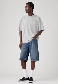 T-shirt cinza em algodão com bolso frontal, combinado com calções de ganga azuis. O conjunto é completado com sapatos pretos sem atacadores e meias brancas.