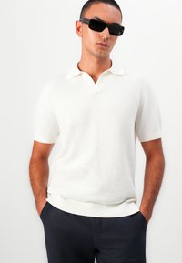 White knitted polo shirt with a v-neckline, short sleeves, and ribbed cuffs. Worn with dark plaid trousers and black rectangular sunglasses.