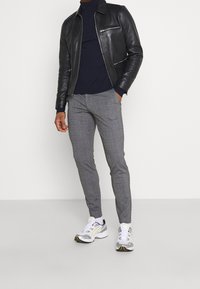 Leren jack, marineblauwe coltrui, grijs gekleurde broek met ruitpatroon en witte sneakers met grijze en gele accenten. Moderne pasvorm, gestructureerde materialen, veelzijdige look.