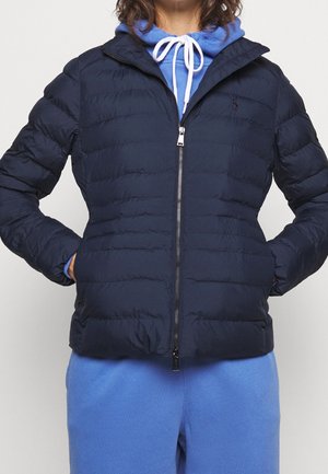Person trägt eine marineblaue Steppjacke mit Reißverschluss über einem blauen Hoodie mit weißen Kordeln, Hände in den Jackentaschen, vor einem schlichten Hintergrund.