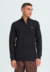 Schwarzer, zipfeliger Strickpullover mit strukturiertem Muster. Mit langen Ärmeln, hohem Kragen und einem kleinen braunen Logo-Patch auf der Brust.