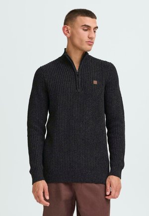 Schwarzer, zipfeliger Strickpullover mit strukturiertem Muster. Mit langen Ärmeln, hohem Kragen und einem kleinen braunen Logo-Patch auf der Brust.