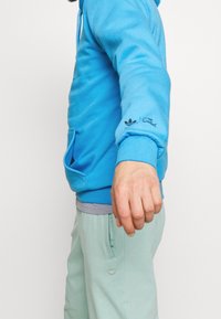 Blå hoodie i mjukt tyg med en framficka. Har en logotryck på ärmen. Matchas med ljusgröna byxor.