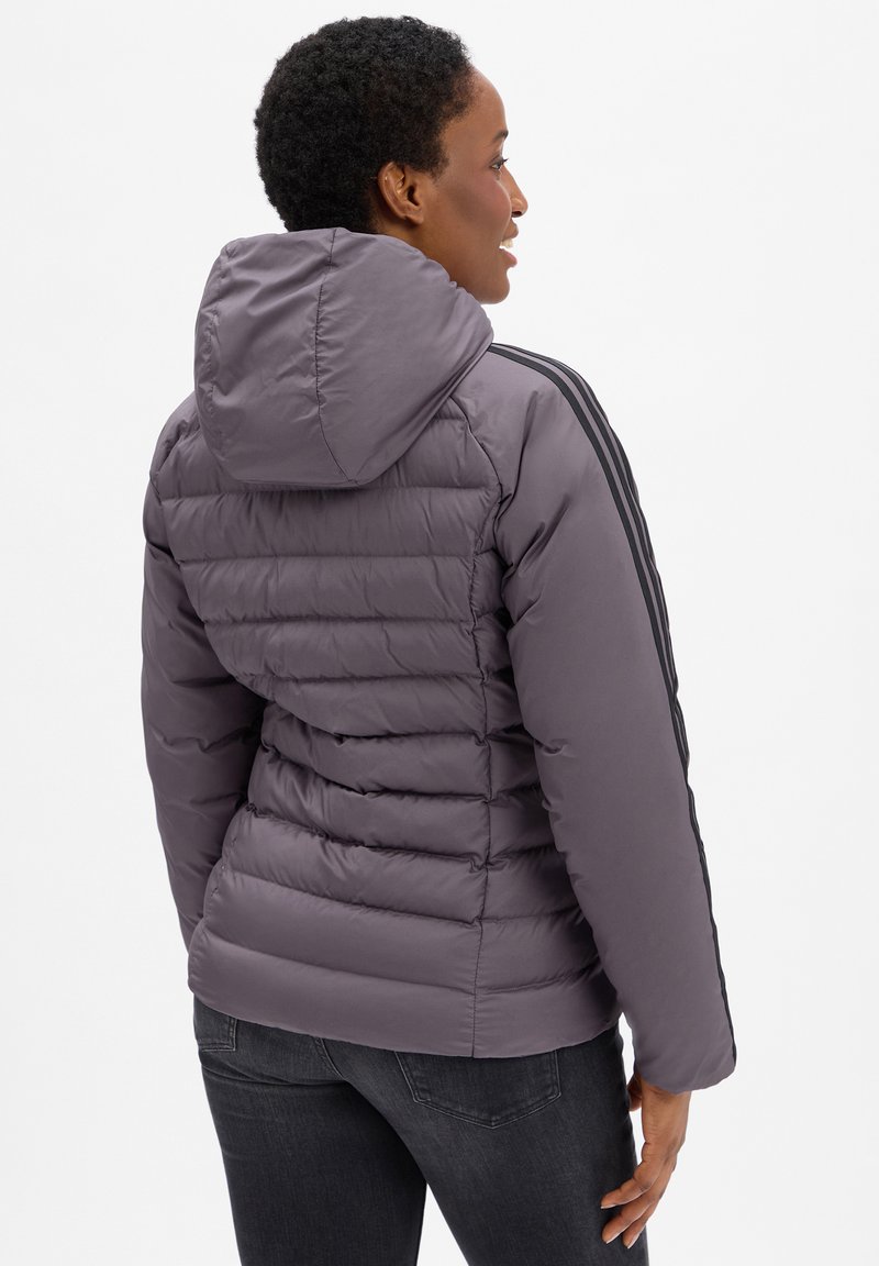 Persona vistiendo una chaqueta acolchada gris con capucha y mangas negras a rayas, girada ligeramente hacia un lado, de pie contra un fondo liso.