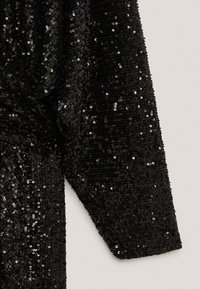 Manche longue d'un vêtement noir recouverte de sequins noirs brillants et densément répartis sur un fond clair uni.