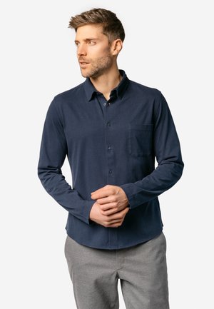 Mann mit kurzen braunen Haaren, der ein marineblaues Hemd mit Knöpfen und graue Hose trägt, steht mit verschränkten Händen und schaut nach links.