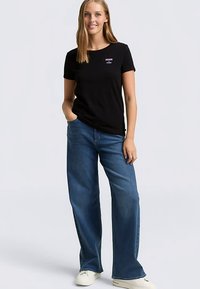 Zwart T-shirt met korte mouwen met witte "DENIM" tekst op de voorkant, gecombineerd met blauwe jeans met wijde pijpen en witte sneakers.