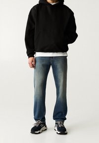 Sudadera negra con capucha y bolsillo tipo canguro, sobre una camisa blanca, combinada con jeans azul claro descoloridos y zapatillas deportivas negras.