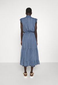 Blauwe maxi-jurk met gelaagde franje, korte mouwen en een aansluitende taille. Lichtgewicht stof met subtiele verticale strepen. Platte sandalen zichtbaar.