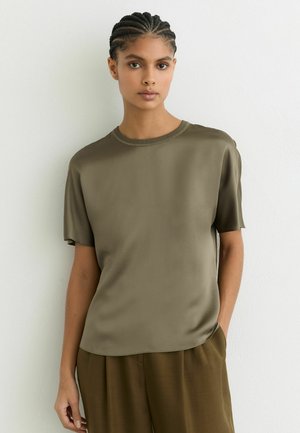 Fonott hajú nő olívazöld rövid ujjú felsőt és hozzáillő nadrágot visel, fehér fal előtt áll, egyik kezét a zsebében tartva.