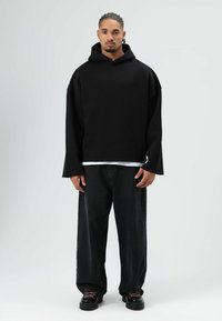 Zwarte oversized hoodie met een brede halslijn, lange mouwen en een matte textuur, gecombineerd met losse zwarte jeans en chunky zwarte sneakers.