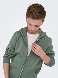 Felpa verde con zip e texture morbida, dotata di cappuccio, tasche laterali e polsini a costine. Indossata sopra una maglietta bianca.