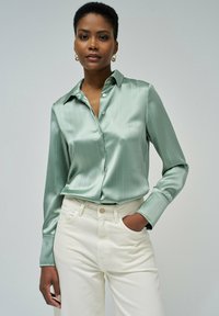Camisa de satén verde claro, de botones, con rayas verticales, mangas largas y cuello, combinada con pantalones blancos de cintura alta.