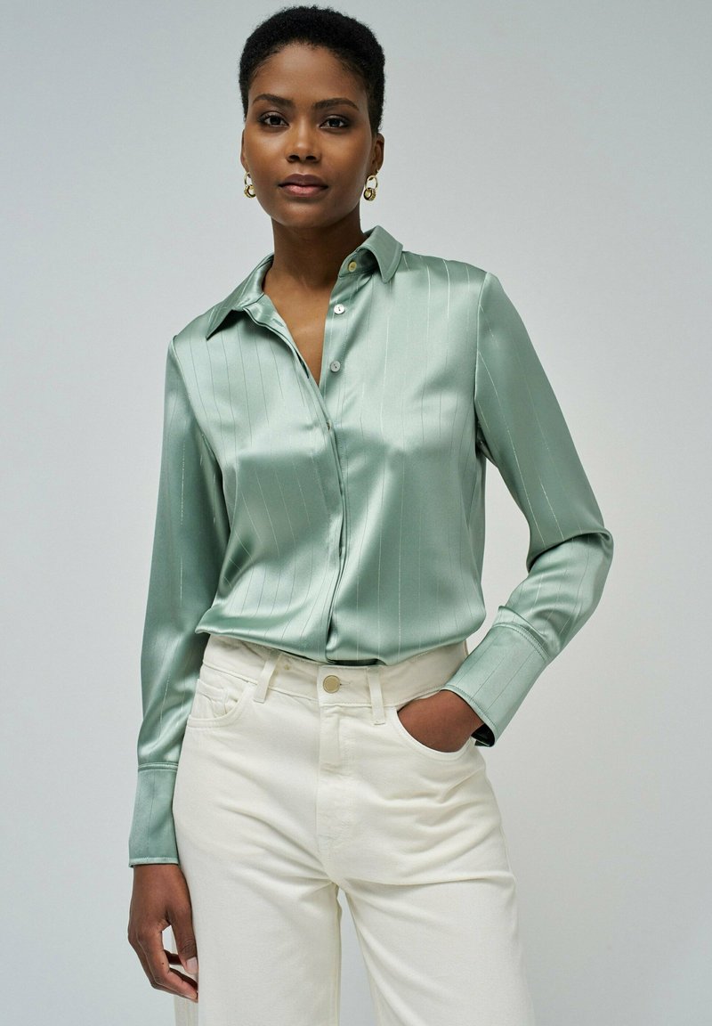 Camisa de satén verde claro, de botones, con rayas verticales, mangas largas y cuello, combinada con pantalones blancos de cintura alta.