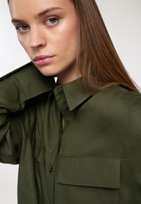 Camicetta verde oliva realizzata in tessuto morbido, con colletto, chiusura a bottoni e due taschini sul petto. Il design prevede una vestibilità rilassata.